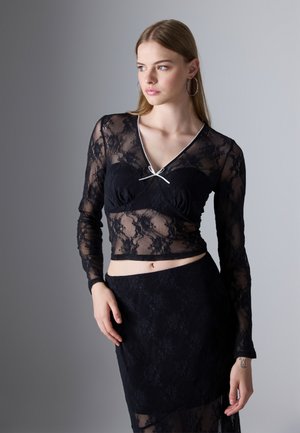 Svart blonde topp med lange ermer, gjennomsiktig stoff, V-hals, og en liten hvit sløyfe detaljer. Stylet med et matchende svart blonde skjørt.