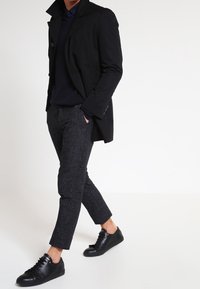 Abrigo negro sobre un suéter azul marino, combinado con pantalones oscuros de textura y zapatillas negras. El abrigo presenta un amplio cuello y detalles de botones.