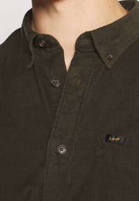 Mörkgrön manchestertröja med knappkrage, försedd med en liten svart etikett med guldfärgad text och bruna knappar på knapplisten.