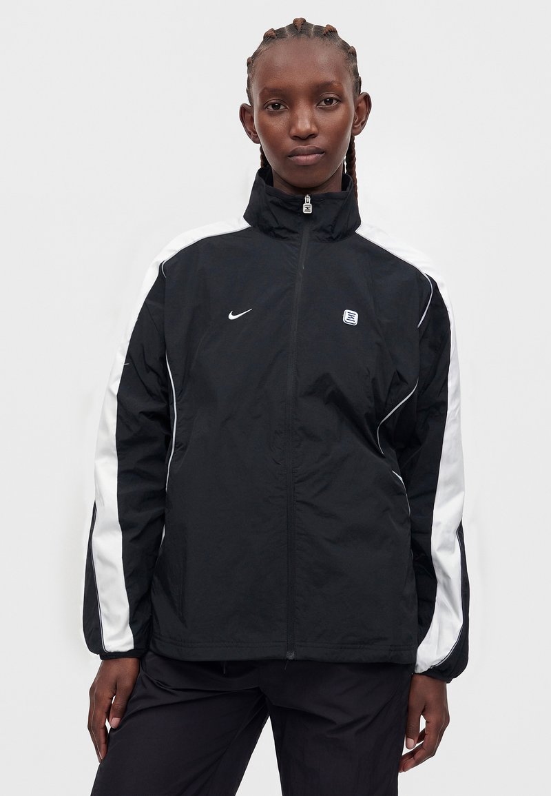 Persoon met gevlochten haar die een zwart-witte Nike jas met rits en hoge kraag draagt tegen een effen grijze achtergrond.