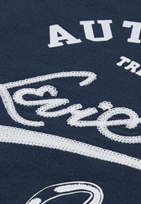 Marinblått tyg med broderad vit text och design. Har texturerad söm och ett böjt vitt band i botten.