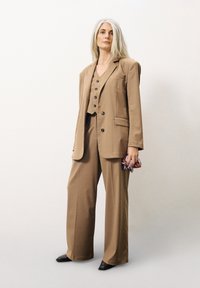 Beige Anzug mit weiten Hosen, strukturiertem Blazer mit einer Brusttasche und einer Weste mit drei Knöpfen, kombiniert mit dunklen Schuhen.