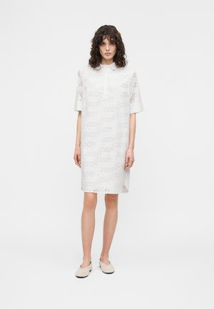 Femme aux cheveux bouclés portant une robe blanche mi-longue à manches courtes avec un motif subtil de texte perforé "JOOP!", debout sur un fond blanc.