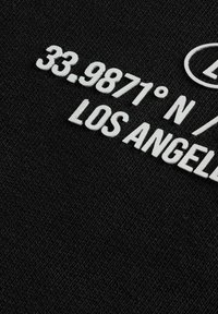 Svart tyg med vit text: "33.9871° N / 118.2437° W LOS ANGELES." Texten har en upphöjd, strukturerad design.