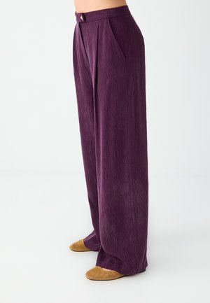 Pantalon violet taille haute à jambes larges avec plis sur le devant, associé à des chaussures plates couleur beige sur une personne sur fond blanc.