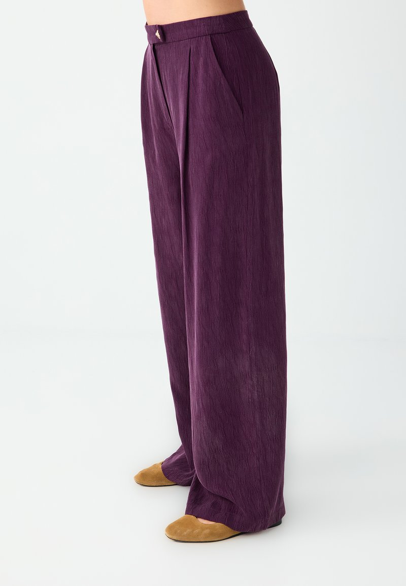 Pantalon violet taille haute à jambes larges avec plis sur le devant, associé à des chaussures plates couleur beige sur une personne sur fond blanc.