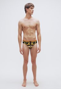 Ανδρικά μαγιό slip σε μαύρο ύφασμα με κίτρινα, χρυσά και λευκά περίτεχνα σχέδια. Εφαρμοστό σχέδιο με χαμηλή μέση.