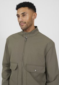 Oliwkowa kurtka z wysokim kołnierzem, zamkiem błyskawicznym z przodu oraz dwoma kieszeniami na piersi z zapięciami na guziki; wykonana z teksturowanego materiału.