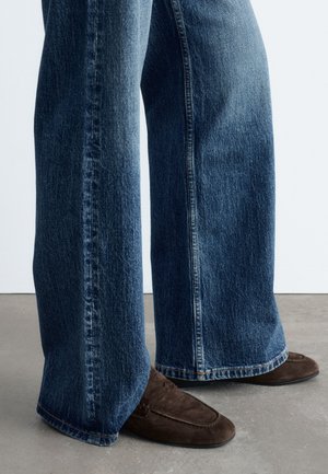 Jambes portant un jean bleu à jambes larges et des mocassins en daim marron, debout sur un sol gris lisse.