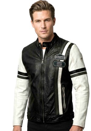 Mann trägt eine schwarz-weiße Lederjacke mit Reißverschluss vorne, gestreiften Ärmeln und Vintage-Patches auf der Brust vor weißem Hintergrund.