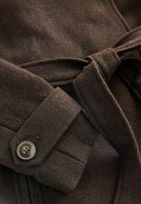Tessuto in lana marrone per un cappotto con vita regolabile. Presenta un dettaglio con bottoni sui polsini e una superficie morbida e strutturata.