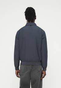 Μπουφάν bomber σε στυλ Navy με καπιτονέ ύφασμα, γιακά, ριμπ μανσέτες και τελείωμα. Μαύρα υφασμάτινα μπαλώματα δημιουργούν διακριτικό μοτίβο. Λεπτομέρεια με κουμπιά στον λαιμό.