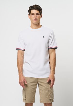 Mężczyzna w białym T-shircie z krótkim rękawem z czerwonym i czarnym obszyciem oraz khaki krótkich spodenkach cargo stoi na tle gładkiego jasnego tła.