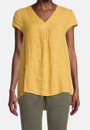 Frau trägt ein locker sitzendes gelbes Kurzarmshirt mit V-Ausschnitt und olivgrüne Hose, steht mit entspannten Armen seitlich an einem einfarbigen Hintergrund.