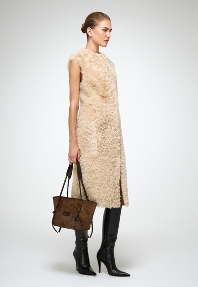 Gilet in shearling beige senza maniche, lungo fino al ginocchio, abbinato a stivali neri con tacco a spillo e che tiene in mano una borsa strutturata marrone.