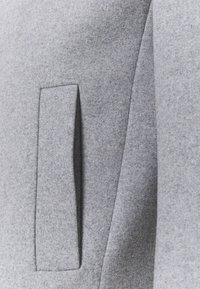 Tela de abrigo de lana gris con un acabado texturizado, que presenta un bolsillo rectangular y una silueta estructurada. Diseño minimalista y tacto suave.