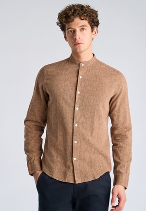 Junger Mann mit lockigem Haar, der ein zugeknöpftes braunes Leinenhemd und dunkle Hosen trägt und vor einem schlichten hellen Hintergrund steht.