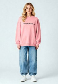 Felpa oversize rosa con testo nero che recita "HO PARLATO DI TE A CHATGPT." Abbinata a jeans ampi blu chiaro e sneakers bianche.