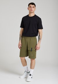 Tricou negru cu mânecă scurtă și buzunar pe piept, asortat cu pantaloni scurți cargo verzi măslinii care au un buzunar lateral mare și un talie elastică. Adidași albi.