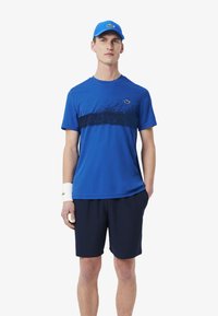 Camiseta atlética azul con una franja azul marino y un patrón moteado, combinada con pantalones cortos azul marino. Incluye una gorra azul y una pulsera blanca.