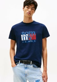 Joven vestido con una camiseta de Tommy Jeans azul marino y jeans rasgados azul claros con un cinturón negro, de pie frente a un fondo blanco liso.