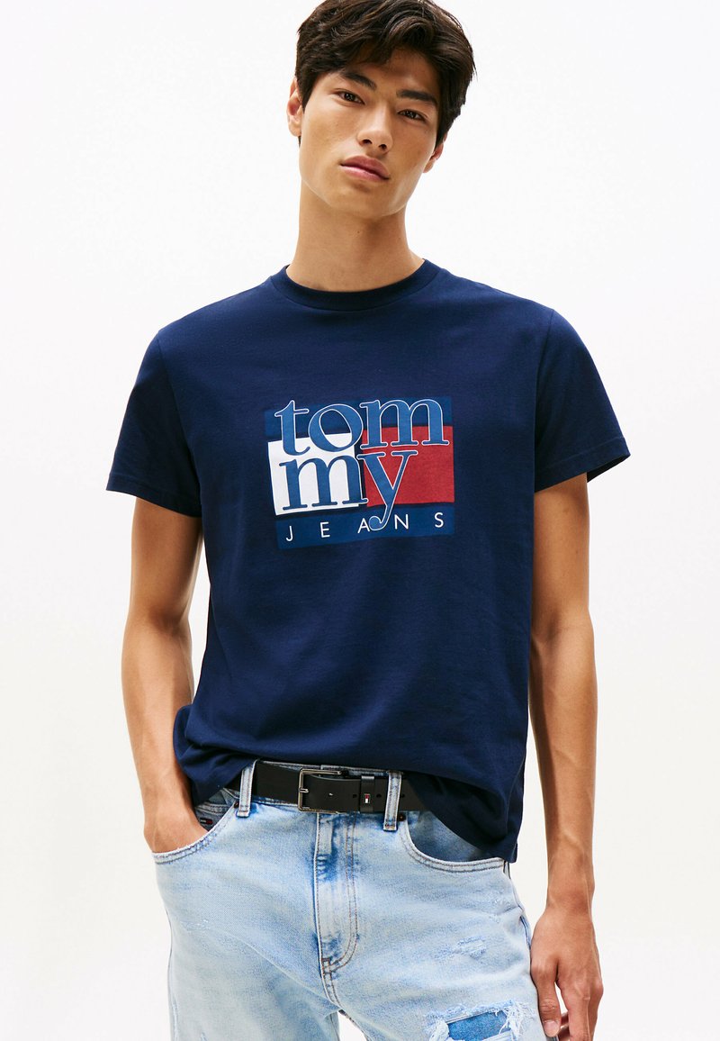 Joven vestido con una camiseta de Tommy Jeans azul marino y jeans rasgados azul claros con un cinturón negro, de pie frente a un fondo blanco liso.