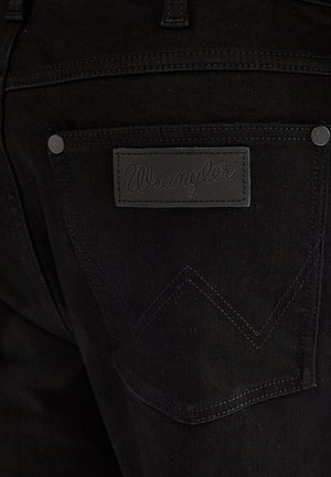 Buzunar spate al blugilor negri Wrangler, cu o etichetă din piele neagră cu numele brandului cusut deasupra buzunarului.