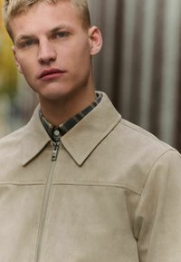 Chaqueta de ante beige con cierre de cremallera, cuello puntiagudo y detalles de costura sutiles. Llevada sobre una camisa de cuadros con detalles de botones.