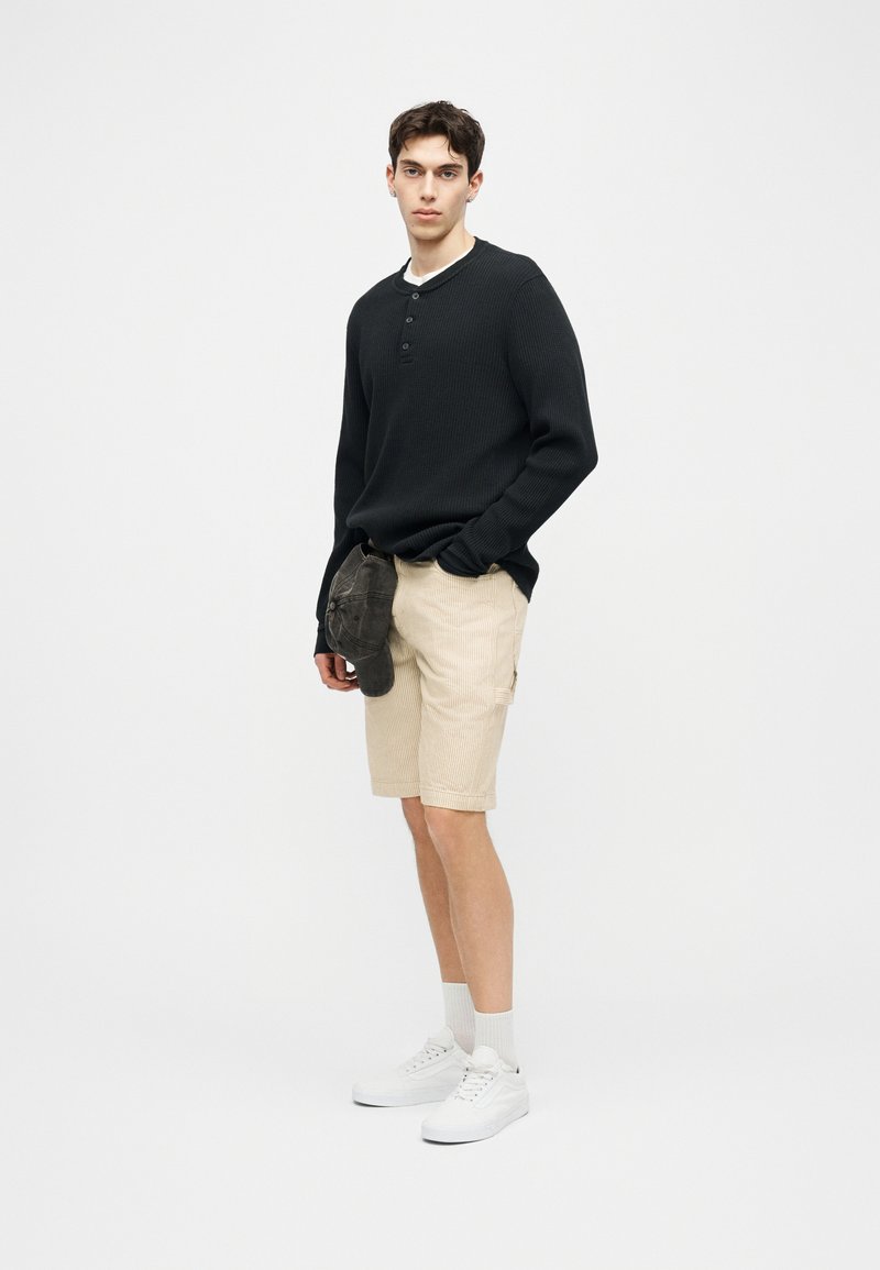 Jonge man draagt een zwarte lange mouw henley trui, beige korte broek, witte sokken en witte sneakers, houdt een donkere pet vast met één hand in zijn zak.
