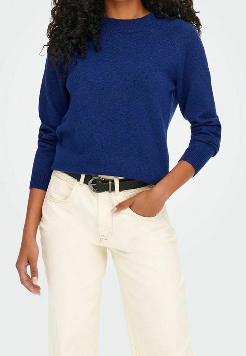 Personne portant un pull à manches longues bleu foncé et un pantalon couleur crème avec une ceinture noire, une main dans la poche, fond blanc.