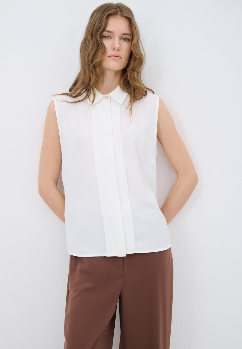 Witte mouwloze blouse met een kraag met knopen, voorkant met plooidetail, gladde stof en subtiele glans, gecombineerd met bruine broek.