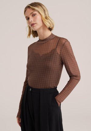 Kobieta o krótkich blond włosach, ubrana w brązową koszulkę w kratę z długim rękawem i czarne spodnie z wysokim stanem, z jedną ręką w kieszeni.