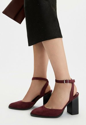 Gambe di donna che indossano scarpe con tacco a blocco in camoscio bordeaux con cinturini alla caviglia e una gonna nera, che tiene una pochette bordeaux coordinata.