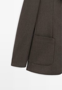 Manica di blazer in tessuto marrone con taglio dritto, caratterizzata da una grande tasca applicata. La texture liscia e le linee pulite evidenziano un design minimalista.