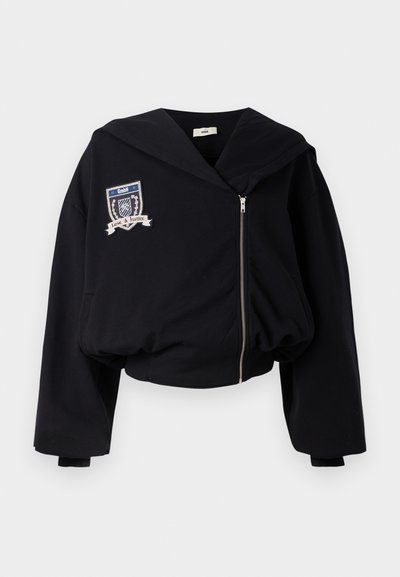 Mustahi hoodie mweusi iliyo katikati na kaptura kubwa, kifungo cha mbele, na alama iliyoshonwa inayoonyesha "GmbH" na "Upendo & Haki." Nyenzo laini ya kitambaa.