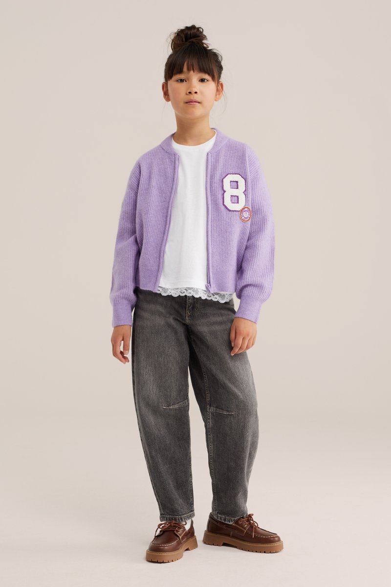 Fille debout portant un cardigan lavande avec un écusson numéro huit, une chemise blanche avec bordure en dentelle, un jean large foncé et des chaussures marron à lacets.