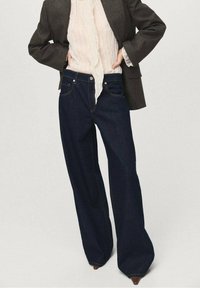 Jeans de pierna ancha azul oscuro con costuras en contraste, que cuentan con cinco bolsillos y un cierre de botón, combinados con una blusa ligera y texturizada y una chaqueta.