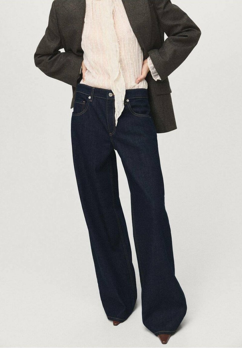 Jeans de pierna ancha azul oscuro con costuras en contraste, que cuentan con cinco bolsillos y un cierre de botón, combinados con una blusa ligera y texturizada y una chaqueta.