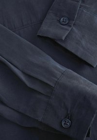 Marineblå skjorte stof med en glat tekstur, med knapmanchetter og en ren syning. Designet inkluderer foldede detaljer og en strømlinet silhuet.