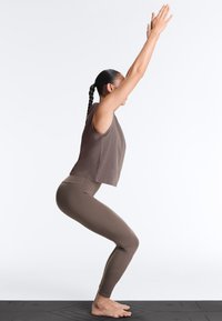 Person i brunt træningstøj, der udfører Utkatasana (stolestilling) med armene hævet, stående barfodet på en sort yogamåtte.