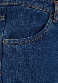 Gros plan sur la poche avant d'un jean en denim bleu avec des coutures orange et des rivets sur la texture du tissu.
