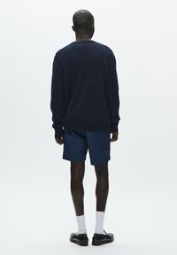 Pull marine avec poignets et ourlet côtelés, associé à un short marine. Le modèle porte des chaussettes blanches et des chaussures noires, vu de dos.