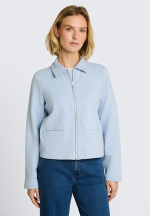 Frau mit kurzen blonden Haaren, die eine hellblaue Jacke mit Reißverschluss und dunkelblaue Jeans vor einem schlichten hellen Hintergrund trägt.