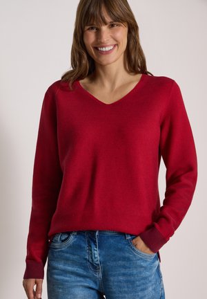 Femme souriante aux cheveux bruns portant un pull rouge à manches longues col en V et un jean bleu, debout avec une main dans la poche devant un fond uni.