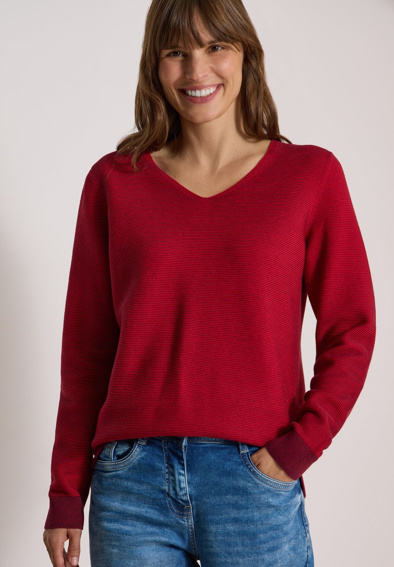Femme souriante aux cheveux bruns portant un pull rouge à manches longues col en V et un jean bleu, debout avec une main dans la poche devant un fond uni.