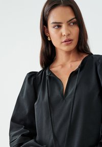 Blusa nera in tessuto morbido e testurizzato, con scollatura a V, maniche a sbuffo e un dettaglio con cordoncino sul davanti.