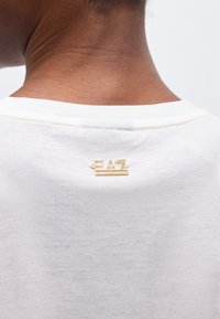 Πίσω μέρος του αυχένα ενός ατόμου που φοράει λευκό πουκάμισο με μικρό χρυσό λογότυπο "EA7" κοντά στον γιακά.