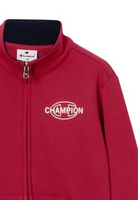 Rode rits-fleece met een zachte stof. Heeft een hoge kraag, wit "CHAMPION" logo en een marineblauwe binnenkraag.