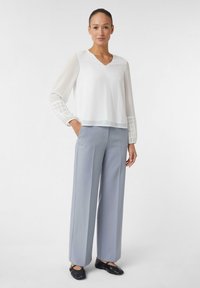 Biała bluzka z dekoltem w serek i przezroczystymi długimi rękawami z siateczkowym wzorem. Do tego jasnoszare, szerokie spodnie oraz czarne buty.