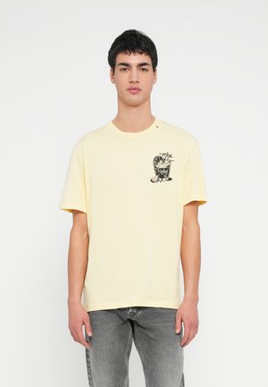 Joven de pie, vistiendo una camiseta amarillo pálido con un gráfico de cóctel negro y jeans grises, contra un fondo blanco liso.